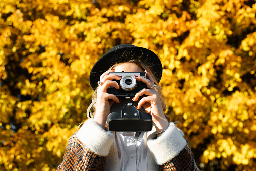 Portrait of young woman hipster photographer in coat and hat taking photo on retro film vintage camera in autumn park outdoors. Selective focus on photographic equipment. Hobby and lifestyle.