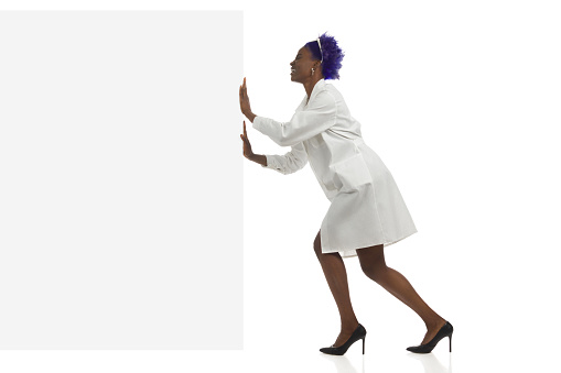 Black woman in lab coat is pushing empty banner and laughing. Side view. Full length.