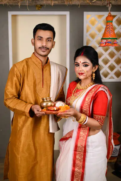 Portrait of Indian men dressed in kurta pajama with beautiful Indian woman wearing traditional Indian saree, gold jewellery and bangles, holding plate of religious offering.