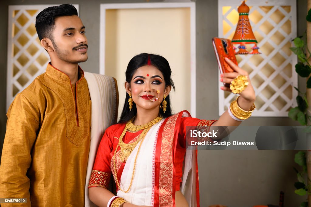 Portrait of Indian men dressed in kurta pajama with beautiful Indian woman wearing traditional Indian saree, gold jewellery and bangles, holding mobile phone taking selfie photo using smartphone. Culture of India Stock Photo