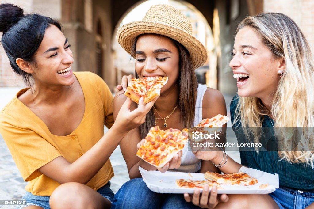Three cheerful multiracial women eating pizza in the street - Happy millennial friends enjoying the weekend together while sightseeing an italian city - Young people lifestyle concept Eating Stock Photo