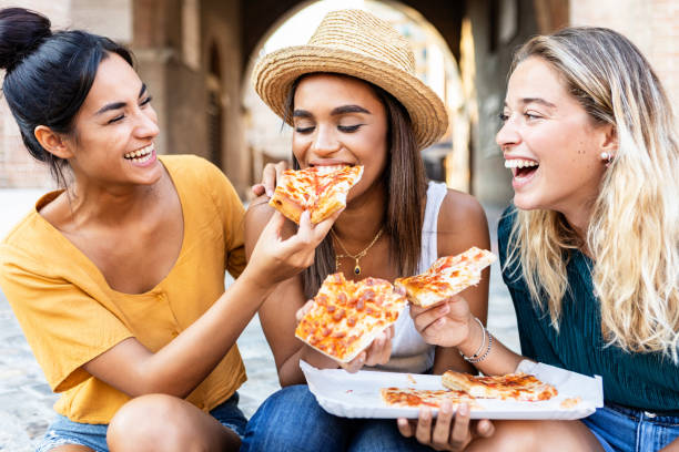 trzy wesołe, wielorasowe kobiety jedzące pizzę na ulicy - szczęśliwi przyjaciele z pokolenia millenialsów cieszący się weekendem razem podczas zwiedzania włoskiego miasta - koncepcja stylu życia młodych ludzi - eating in zdjęcia i obrazy z banku zdjęć