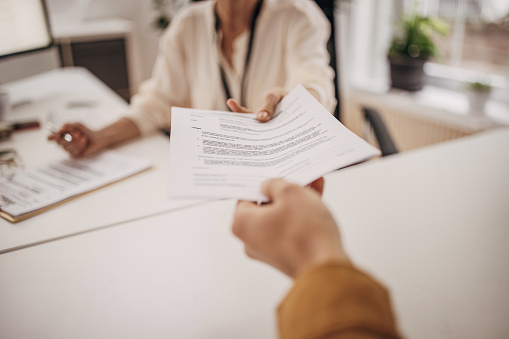 Two people, mature businesswoman sitting at her desk in office, giving documents to young male colleague.