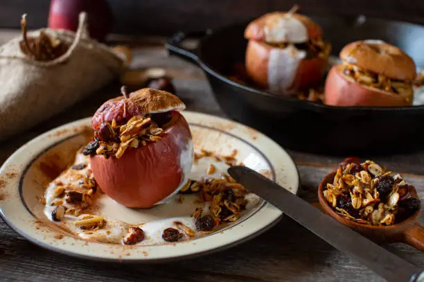 Delicious warm and healthy breakfast for autumn and winter season with oven baked apple filled with homemade granola and topped with yogurt and cinnamon. Served on a rustic plate on wooden table