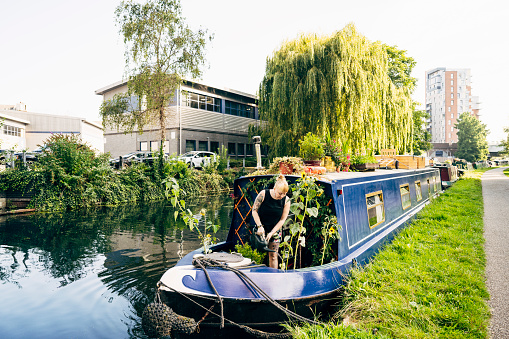 Front view of British woman in casual summer attire tending to garden on bow deck of 57 foot narrowboat moored along grassy embankment.