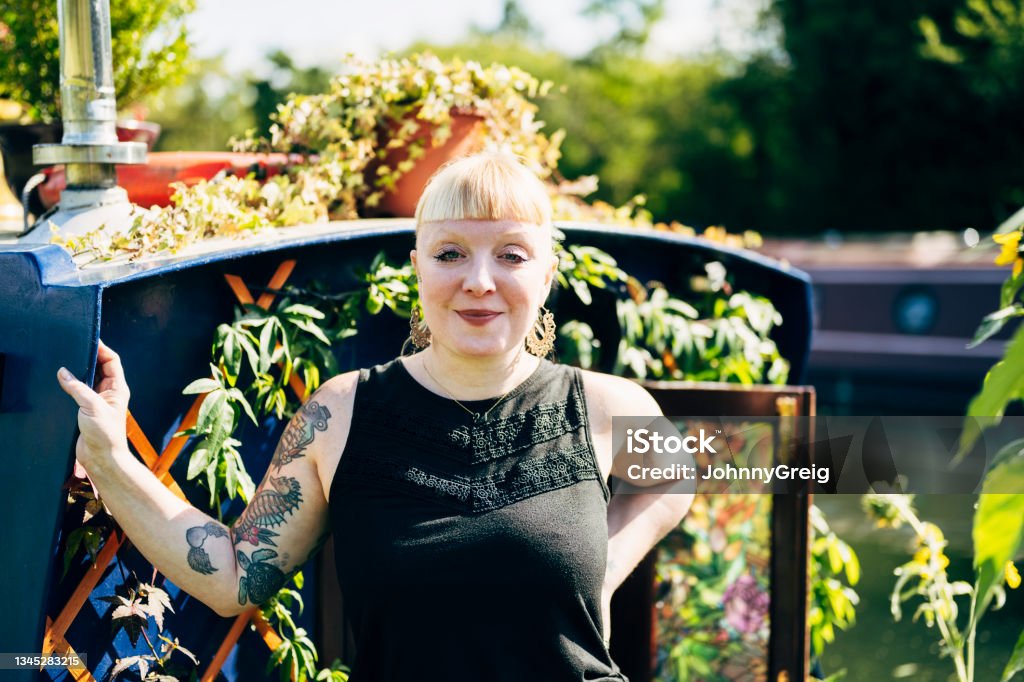 Portrait of British woman and garden onboard narrowboat Waist-up view of blond late 30s woman in casual summer attire standing amidst vining plants and flowers growing on sunny bow deck and smiling at camera. Outdoors Stock Photo