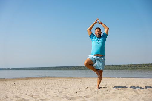 Mid adult man in blue shorts and T-shirt doing yoga exercise on sandy beach, arms raised and clasped, standing on one leg, the other bent inwards, tree pose Vrksasana