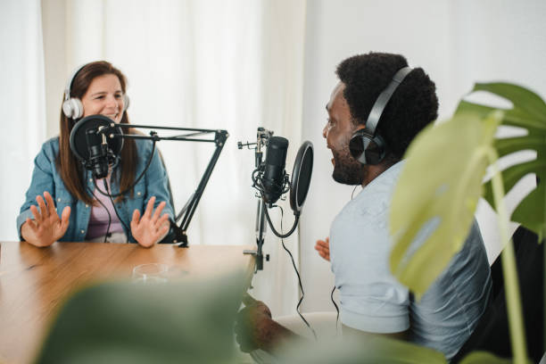 Diverse colleagues recording podcast and talking in microphones Content multiracial male and female coworkers speaking in mics while looking at each other and recoding podcast in modern studio podcast stock pictures, royalty-free photos & images