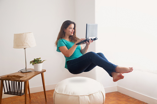 Charming girl casually dressed holding laptop levitating in the air. Working from home can be a dream concept. Floating air and working. Indoors