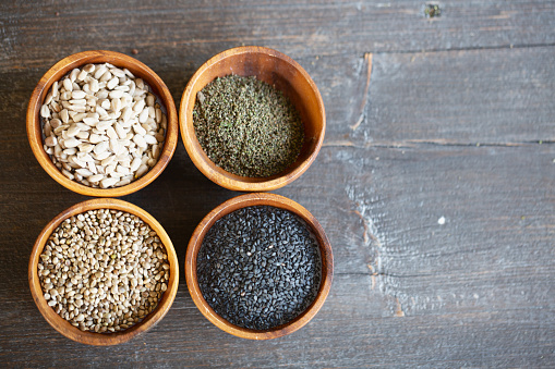 A variety of plant based proteins: seeds in wooden bowls on a dark wooden background, seen from above. These vegan proteins are: sesame seeds, sunflower seeds, hemp seeds, nettle seeds, r