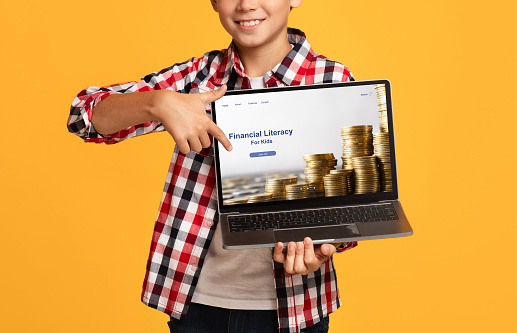 Cropped view of happy boy pointing at laptop screen with Financial Literacy For Kids educational website, orange studio background. Online economy school, money management course