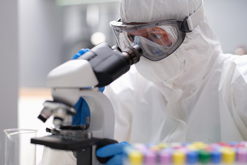 Scientist chemist in protective anti-plague suit looking through microscope in laboratory. Scientific research on coronavirus infection concept