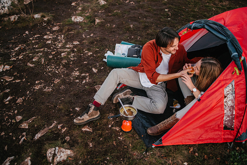 Smiling young couple eating and talking in front of their red tent on a hiking trip. Man is feeding his girlfriend