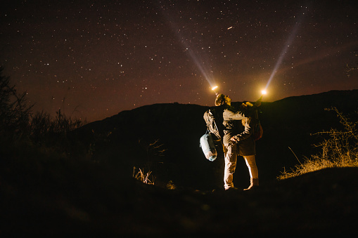 A couple of hikers hugging and looking at starry sky, pointing lamps at a sky