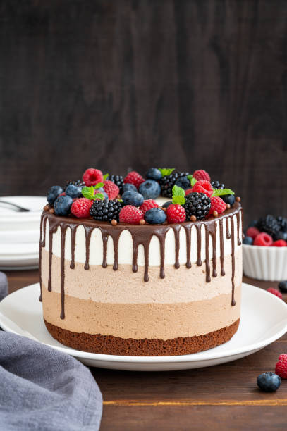 istock Three chocolate mousse cake with chocolate glaze, blueberries, raspberries and blackberries on top on a white plate on a dark wooden background. Cake with three layers of different taste. Copy space. 1345313582