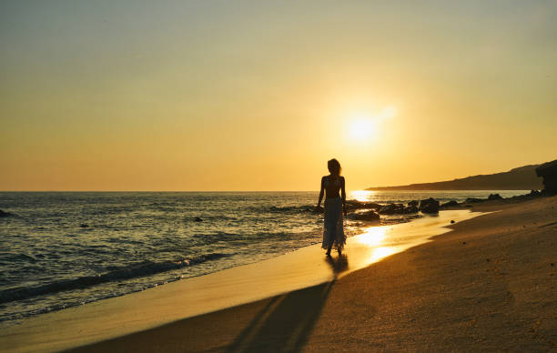 日没時に砂浜に沿って歩く気ままな若い女性 - deep focus 写真 ストックフォトと画像