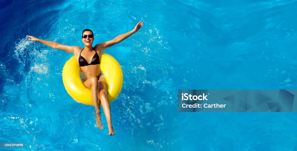 Happy laughing young woman enjoying the water park Summer vacation. Beautiful slim young woman enjoying an aqua park floating in a yellow big ring on sparkling blue pool while smiling at camera, panorama top view with copy space Swimming Pool Stock Photo