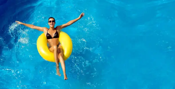 Summer vacation. Beautiful slim young woman enjoying an aqua park floating in a yellow big ring on sparkling blue pool while smiling at camera, panorama top view with copy space