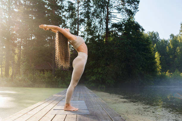 A woman practicing yoga performs an exercise, back bend, Hasta Uttanasana pose, stands on a wooden bridge on a sunny warm morning in a park near a pond