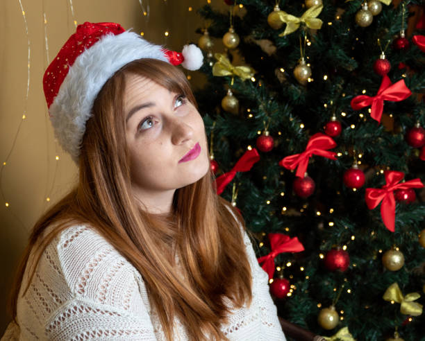 istock A red-haired girl in a New Year's hat with a thoughtful expression on her face. Decorated Christmas tree on a blurry background. Selective focus. 1345350502