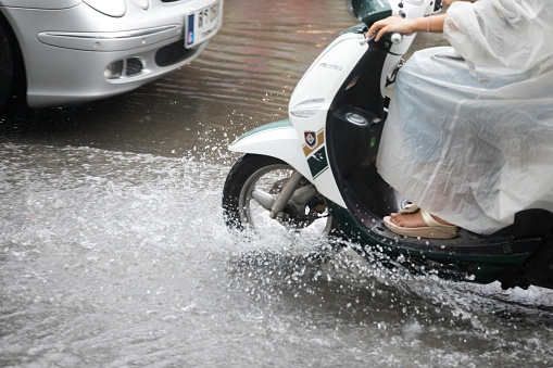 Thai woman in rain cape on motorcycle passing flooded street in Bangkok during rainy season