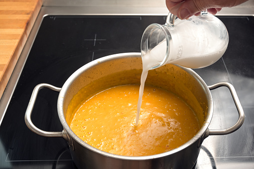 Pouring coconut milk or cream in a vegetable soup from red kuri squash or Hokkaido pumpkin in a steel pot on a black stove, cooking an autumn meal, copy space, selected focus, narrow depth of field