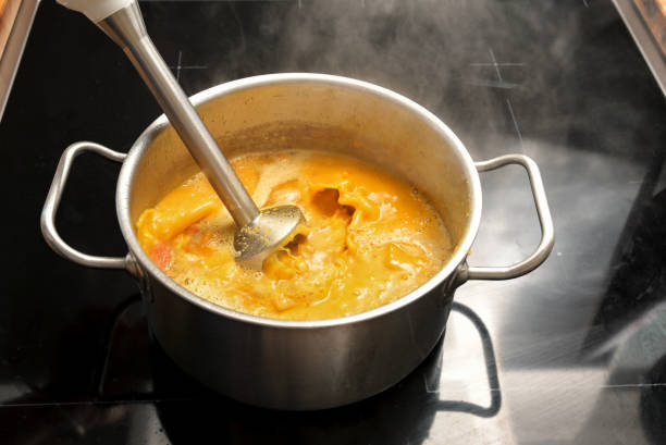 Vegetarian vegetable soup from Hokkaido or red kuri squash is pureed with an immersion blender in a steel pot on a black stove, cooking in autumn, copy space, selected focus, narrow depth of field stock photo