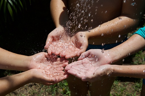 Clean water falls from a plant watering can into children's hands to cool a sunny and hot day.