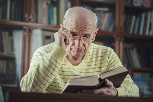 Aged male person holding glasses with hand and reading book sitting at table indoor