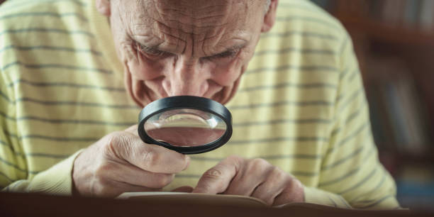 istock Elderly man with magnifying glass in hand reading book on table 1345371664