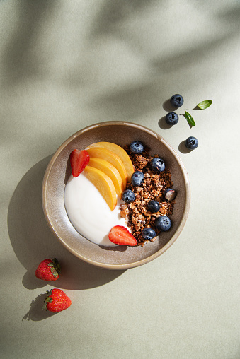 Delicious natural yogurt with homemade granola; peach, berries  in a bowl on a green background with morning shadows. Healthy and nutritious breakfast. Diet food. Top view and copy space. Vertical