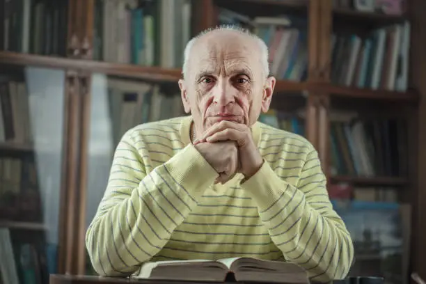 Photo of Senior male author sitting at table with book and holds hands under head.