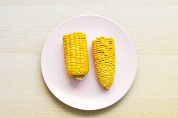 An ear of hot boiled yellow corn is broken in half, it has just been boiled and then sprinkled with salt and greased with butter, lies on a pink round flat plate on a brown table, top view, copy space.