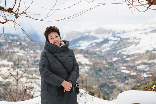 Portrait of senior beautiful woman in long grey coat with splendid snow-covered mountains view in background. Winter vacation