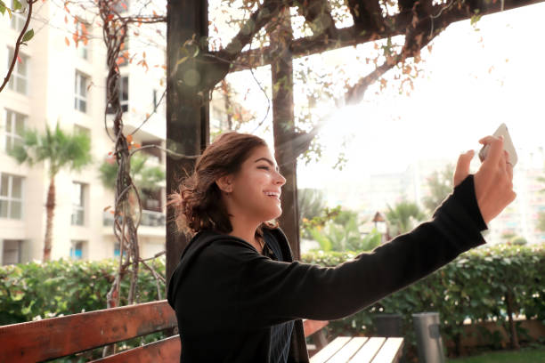 portrait of  beautiful young woman taking selfie with cellphone - using phone garden bench imagens e fotografias de stock