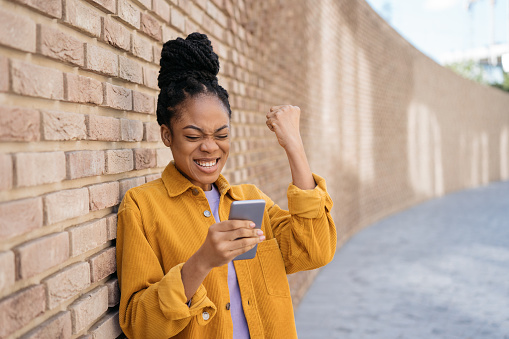 Mujer afroamericana emocional que usa el teléfono móvil leyendo buenas noticias sobre el resultado del examen. Hermosa y feliz mujer gana éxito de celebración de lotería en línea photo