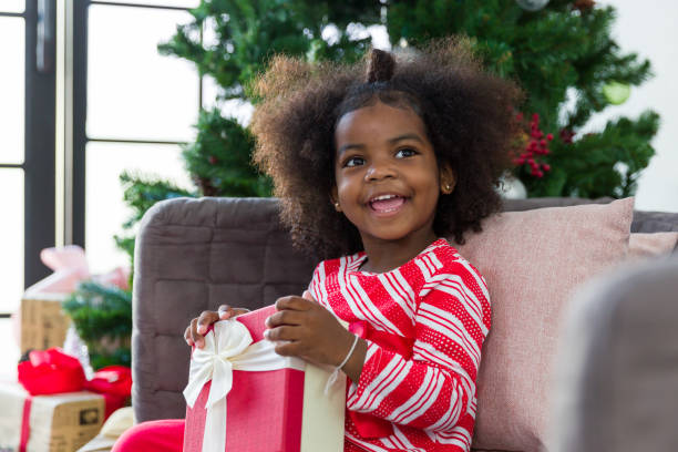 heureuse mignonne petite fille afro-américaine souriante avec un coffret cadeau de noël ou un cadeau cadeau à noël à la maison. fêtes de noël, joyeux noël et joyeuses fêtes - christmas child baby surprise photos et images de collection