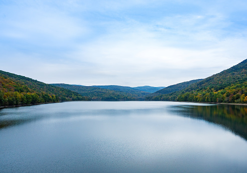 Beautiful calm lake surrounded by forest. Colors of the leaves starting to change with the fall season, and reflections in the water. Blue sky, copy space. Pepacton Reservoir, Catskill Mountains, New York State.