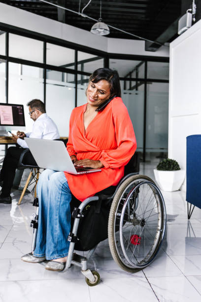 istock Latin transgender woman sitting in wheelchair using computer while talking on the phone in office in Mexico 1345432179