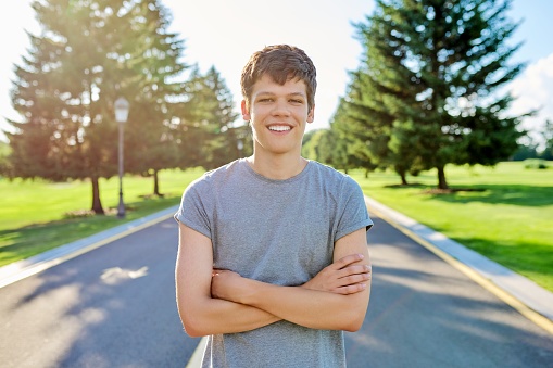 Portrait of a guy 16, 17 years old looking at the camera. Smiling teenager with crossed arms, on the road in the park, summer sunset background