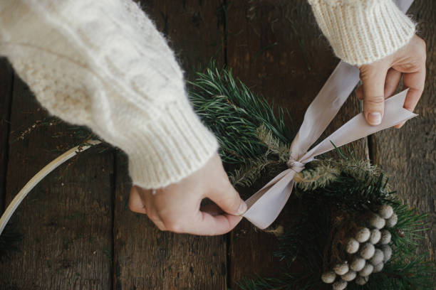 istock Making modern boho christmas wreath. Hands tying ribbon on xmas wreath with fir branches, brunia herb, round wooden hoop on rustic table. Atmospheric moody image. Winter holidays preparation 1345444045