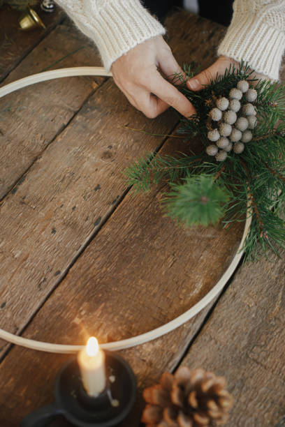 istock Making modern minimalistic christmas wreath. Hands making wreath on rustic table with fir branches, brunia herb, ribbons, round wooden hoop, thread, candle. Atmospheric moody image 1345444819