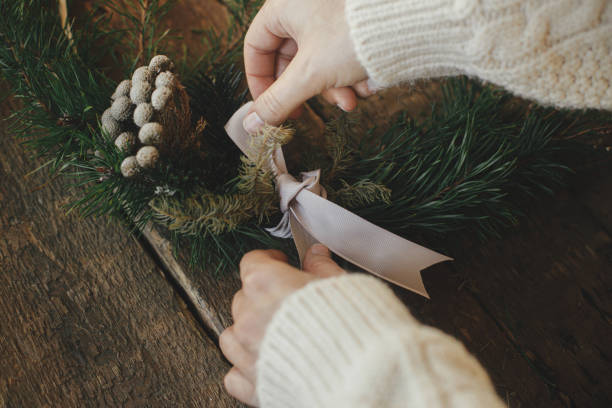 istock Hands tying ribbon and making modern christmas wreath on rustic table with fir branches, brunia herb, round wooden hoop. Atmospheric moody image. Winter holidays preparation close up 1345444951