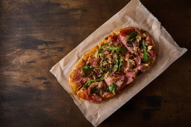 Rectangular Roman pizza with prosciutto ham, tomatoes, mozzarella, mushrooms and arugula on wooden background, flat lay. Copy space for text