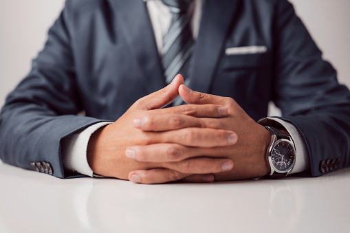 Clasped hands of a businessman in a formal suit sitting at a work or meeting table. An attentive professional taking part in interviews.