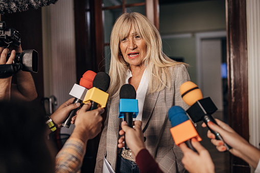Group of people, female politician confronted by journalists with microphones.
