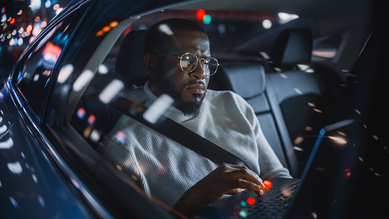 Stylish Black Man in Glasses is Commuting Home in a Backseat of a Taxi at Night. Male Using Laptop Computer and Looking Out of Window while in a Car in Urban City Street with Working Neon Signs.