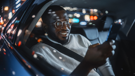 Stylish Black Man in Glasses is Commuting Home in a Backseat of a Taxi at Night. Handsome Male Using Smartphone and Looking Out of Window while in a Car in Urban City Street with Working Neon Signs.