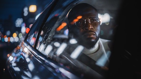 Stylish Black Man in Glasses is Commuting Home in a Backseat of a Taxi on a Rainy Night. Handsome Male Passenger Looking Out of Window while in a Car in Urban City Street with Working Neon Signs.