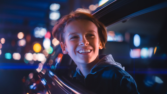 Excited Young Boy is Sitting on Backseat of a Car, Commuting Home at Night. Looking Out of the Window with Amazement of How Beautiful is the City Street with Working Neon Signs.